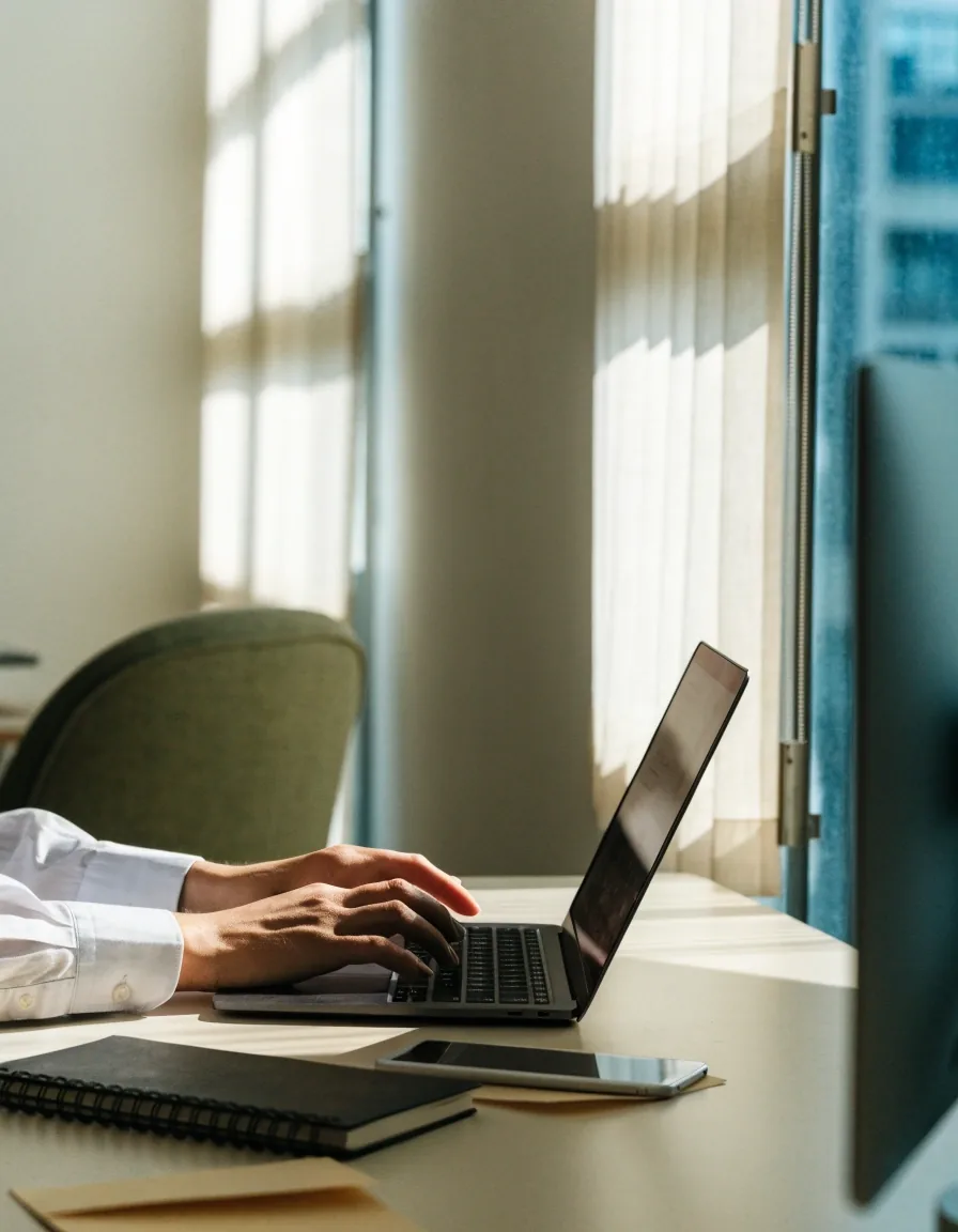 Hands at a laptop on a clean desk in a bright consultancy office, notebook and phone set aside